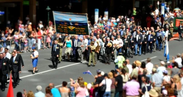 The banner they carry names the dead. The people beneath it are still here. That gap between the two is what ANZAC Day is about.