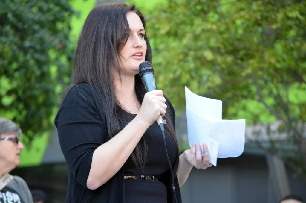 A speaker reading from notes at an outdoor rally looks like testimony. The notes matter — they mean someone prepared, someone thought carefully about what to say.