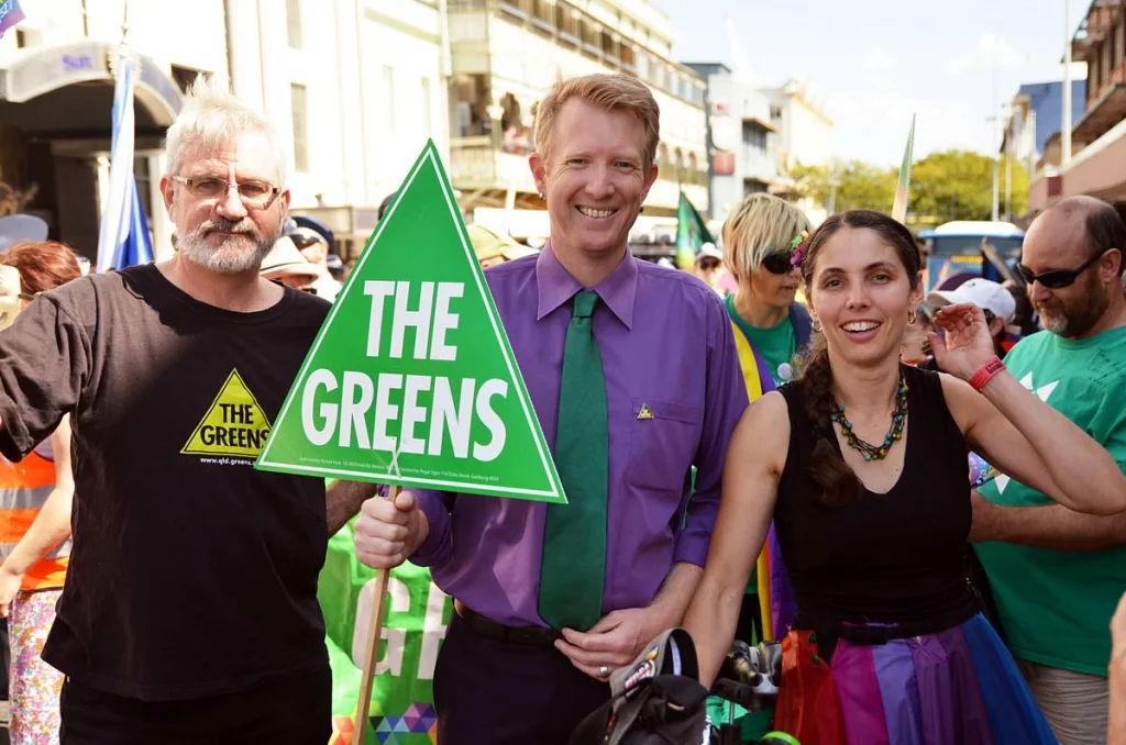 The Greens triangle sign in green, held by someone in a purple shirt at a pride parade — an accidental, perfect colour combination.