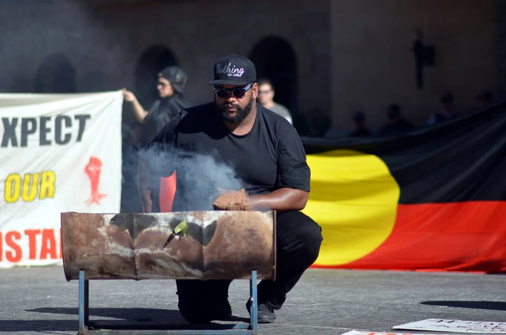He crouched over the smoking barrel with the Aboriginal flag behind him. Ceremony and protest occupying the same space without contradiction.