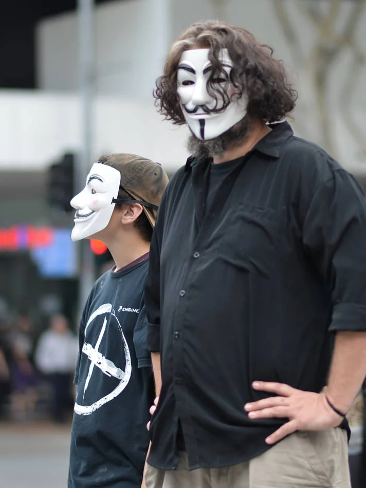 A father and child in matching Guy Fawkes masks, looking up at the speakers together. The masks make identification impossible and the image universal.
