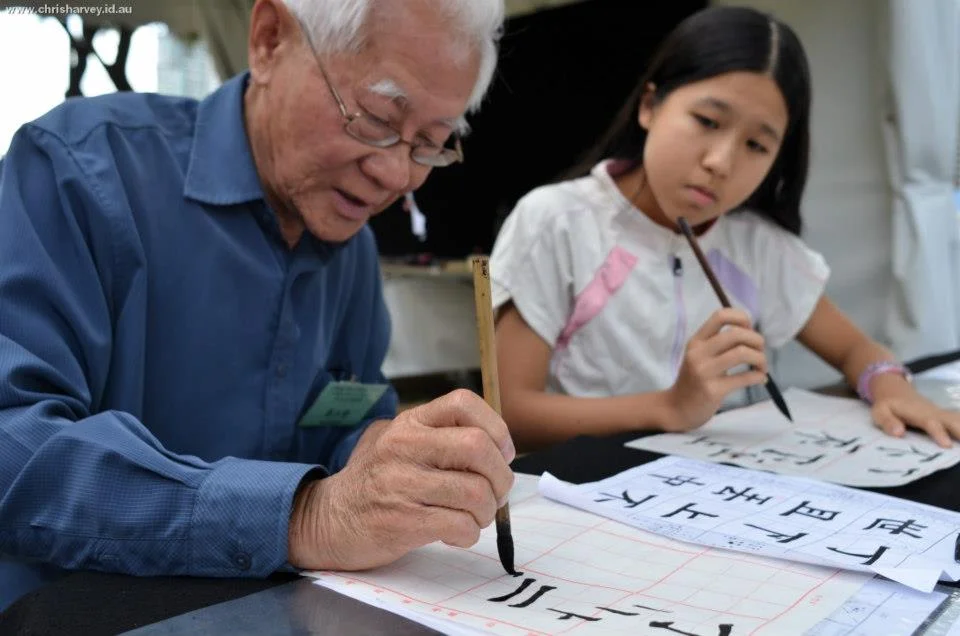 An elder and a child, both holding brushes, both focused. The gap in their hands is sixty years. The technique is the same.