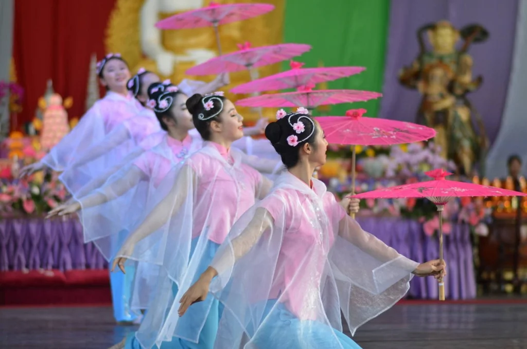 Four dancers in a diagonal line, each holding a pink parasol at the same angle. Synchrony this precise takes years to achieve and half a second to photograph.