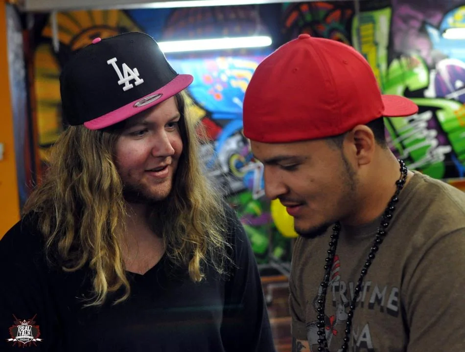 Two rappers against a graffiti mural, mid-conversation before their battle. The wall does half the composition work.
