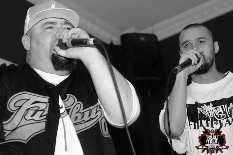 Two microphones, two performers, one frame. The mics are halfway to their mouths and the round hasn't started yet. That tension is the photograph.