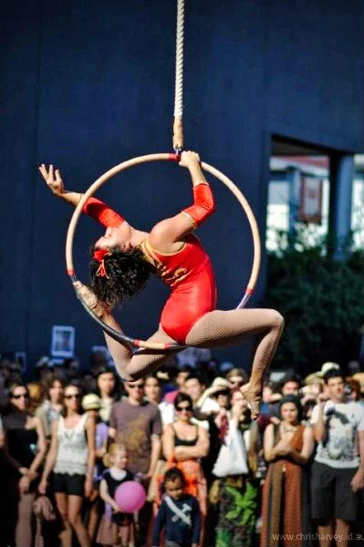 Aerial hoop against a blue building and bright sky. The performer's body makes a shape that only exists at one point in the rotation.