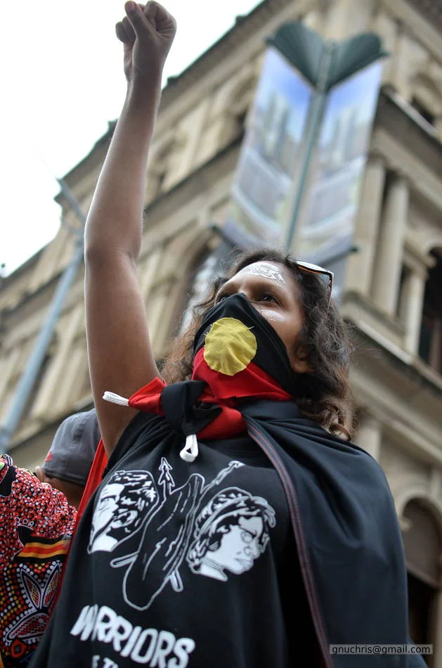 Fist up, flag across her face, the old Brisbane city hall behind her. The frame contains two very different versions of this country.