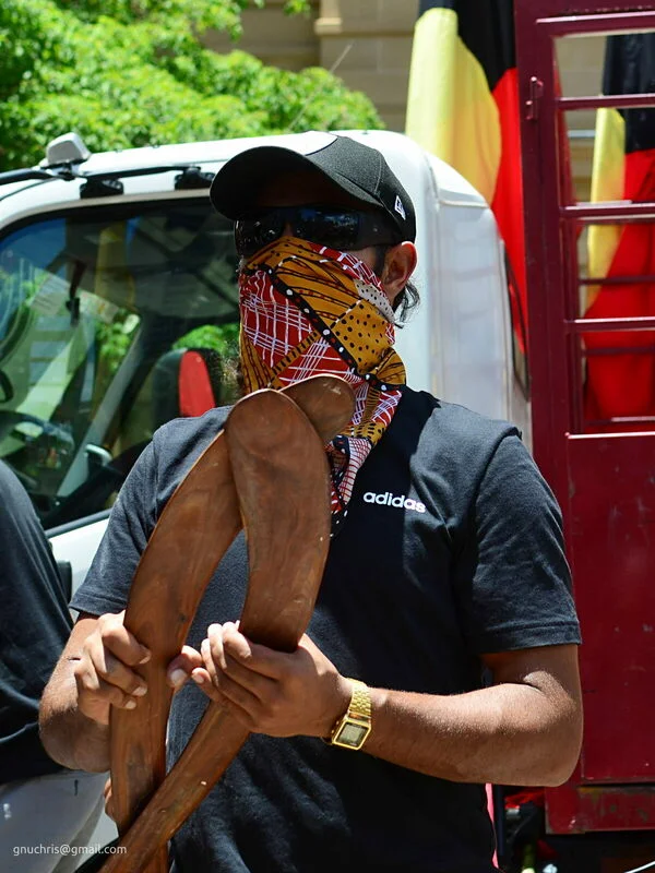 Boomerang in hand, face covered in Aboriginal art fabric. The flags are directly behind him. Everything in the frame belongs there.