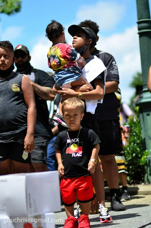 The boy in the Black Flag shirt is watching everything with the particular seriousness of a child who understands what he is part of.