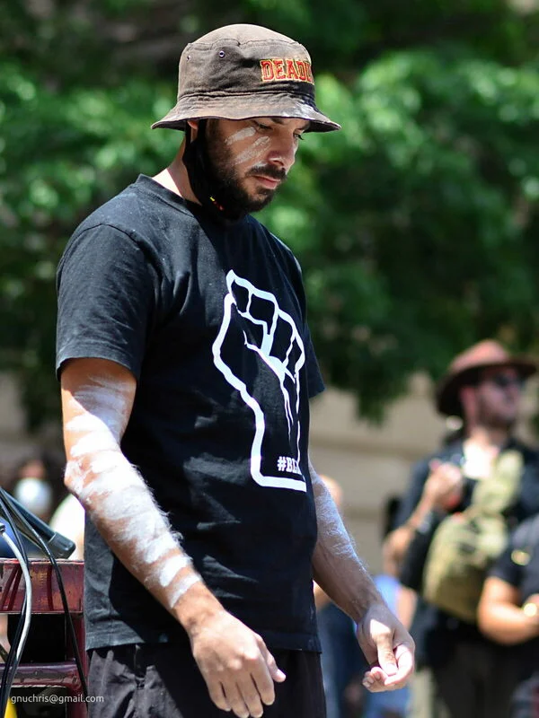 White clay on his arms and face, eyes down, the march moving around him. He is completely present and completely contained.