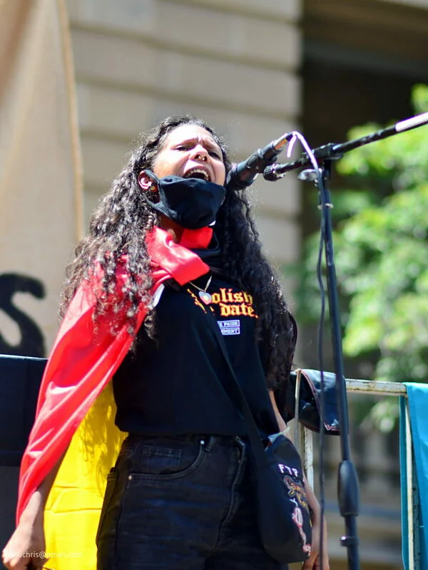 Her voice carries to the back of the crowd. The Aboriginal flag is around her shoulders and she is not done yet.