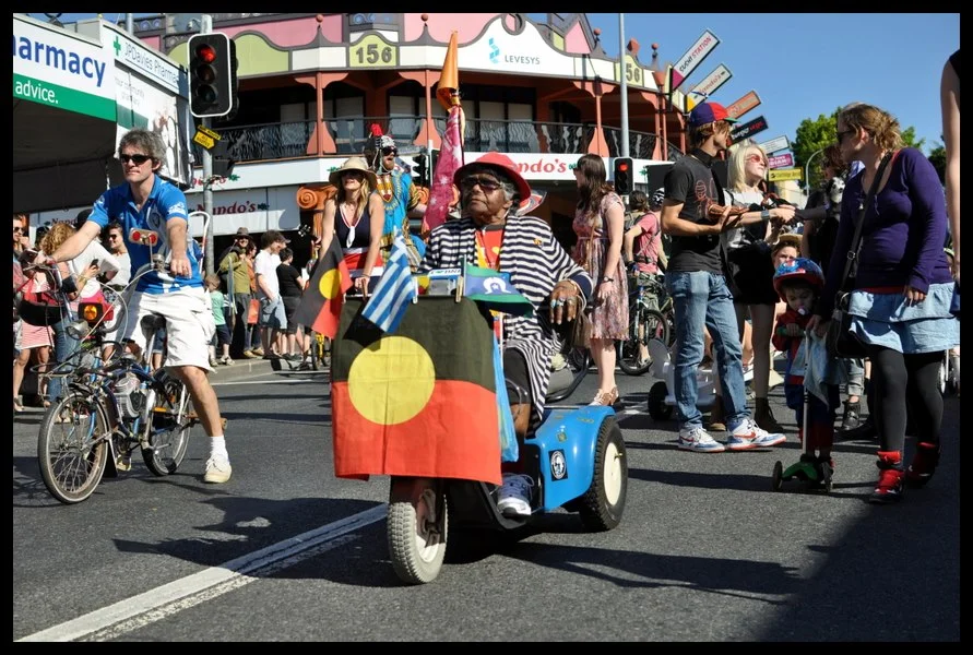 An elder on a mobility scooter, Aboriginal flag attached to the front, moving through the crowd at the intersection. She is in no hurry. Nobody expects her to be.