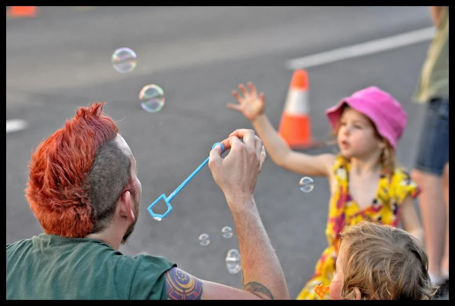 Red mohawk, bubble wand, child reaching forward with both hands. The street is closed to traffic and all of this is possible.