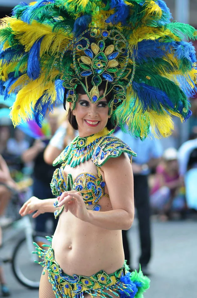 Peacock feathers in green and blue and gold, the face turned slightly away, a smile at something in the crowd. The costume is architectural.