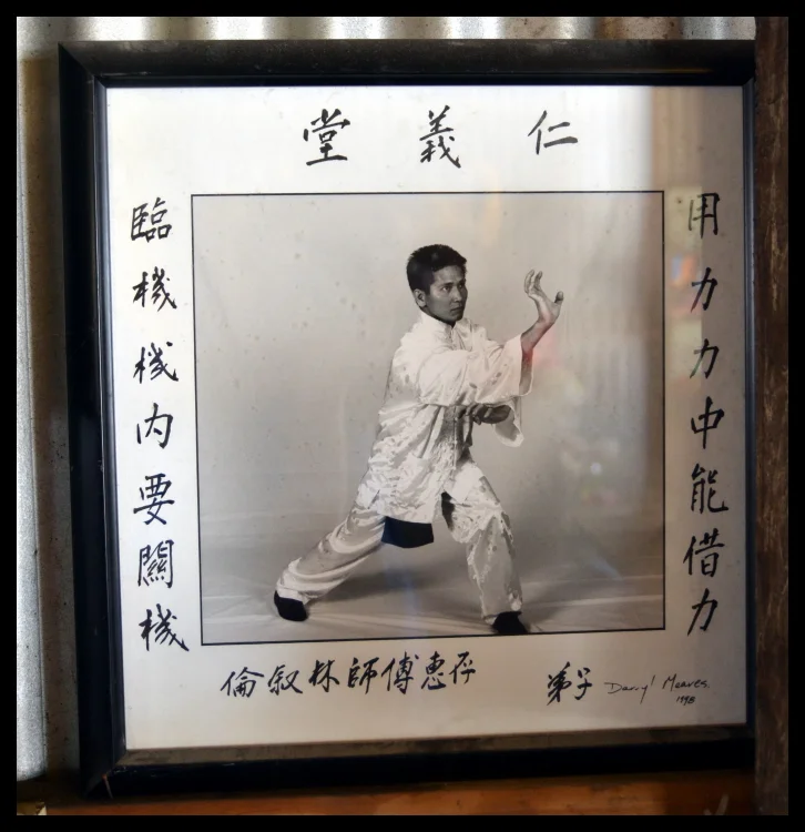 A framed photograph on the school wall — the master in stance, the characters beside him. The lineage is on the wall before the performance begins.