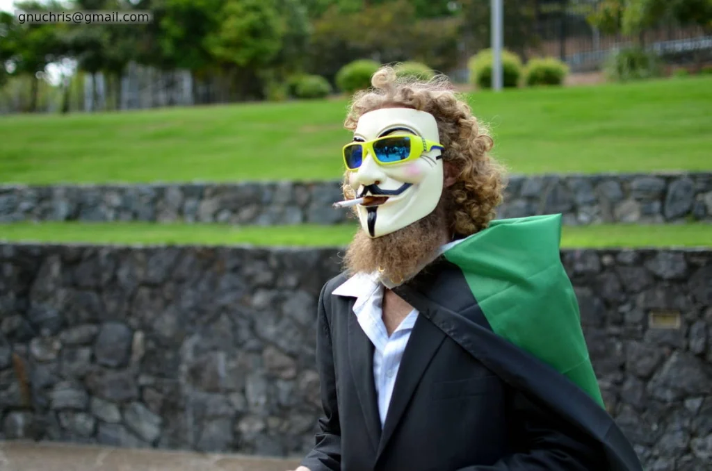 Curly hair, beard extending below the mask, sunglasses over the eyeholes, Anonymous flag over his shoulder. He is fully committed to the aesthetic.
