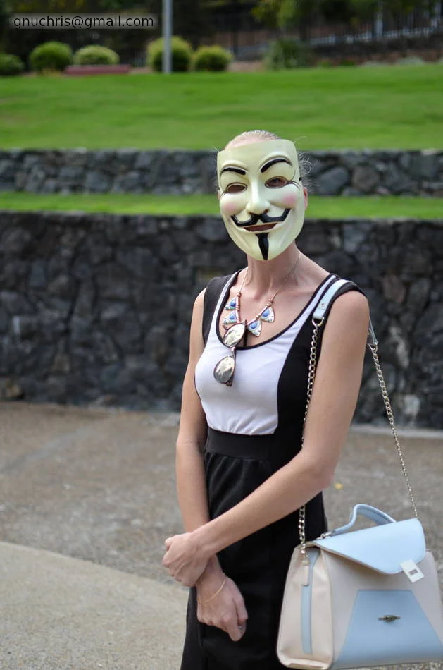 The Guy Fawkes mask with a statement necklace and a pale blue handbag. The costume is the mask — everything else reads normally.