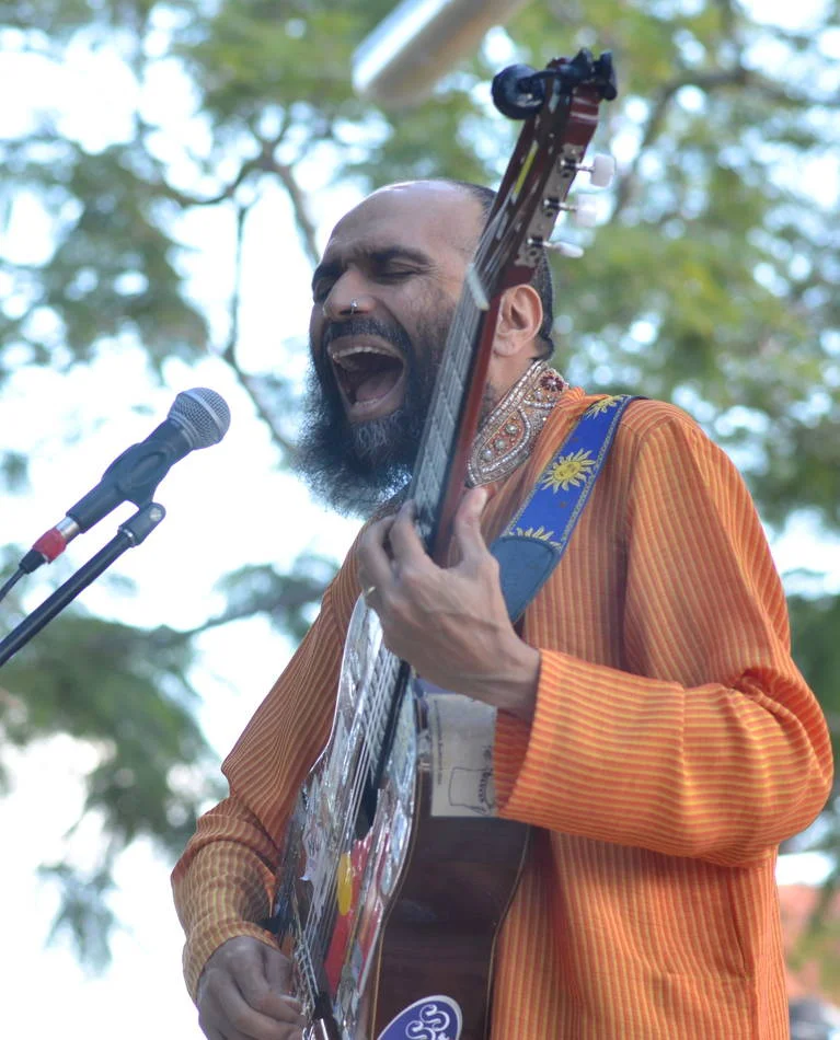 Eyes closed, mouth open, playing something that matters to him. The decorated guitar strap, the orange kurta. He is completely in it.