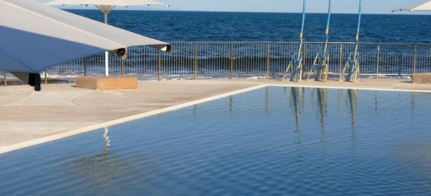 The ocean pool at low tide, the sea just beyond the rail. The water is completely still. The reflection holds everything.