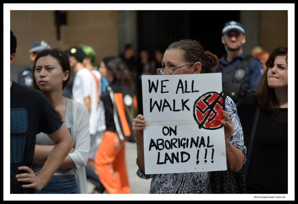 The simplest signs are the most photographable. Four words and a crossed-out symbol is not a slogan — it is a fact with a red pen through a lie.